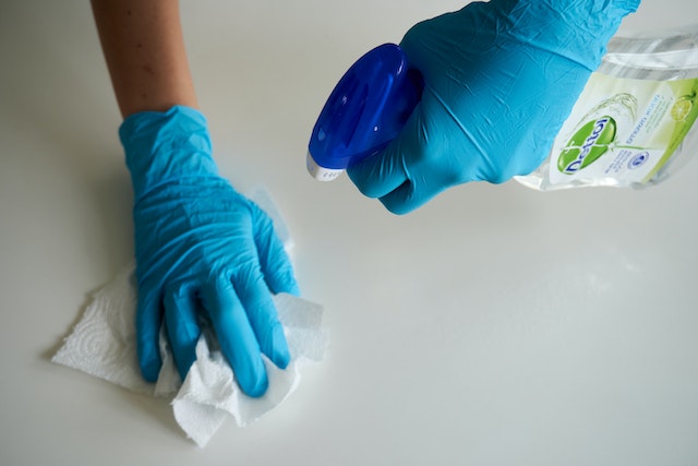 Blue gloved hands spraying a bottle of cleaner and wiping down a counter with a paper towel
